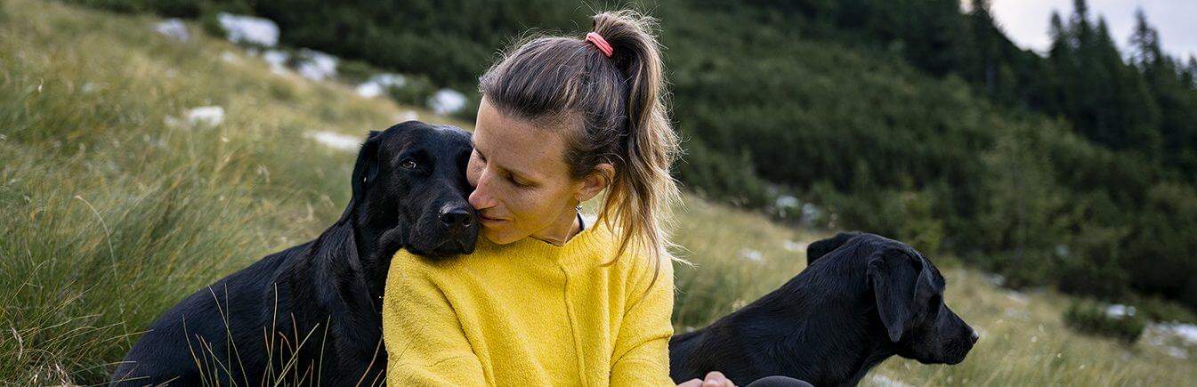 Young female owner of two beautfiul purebred black labrador retriever dogs sitting in green grass up the hill with one of her dogs leaning her head on the women shoulder in a loving gesture.
